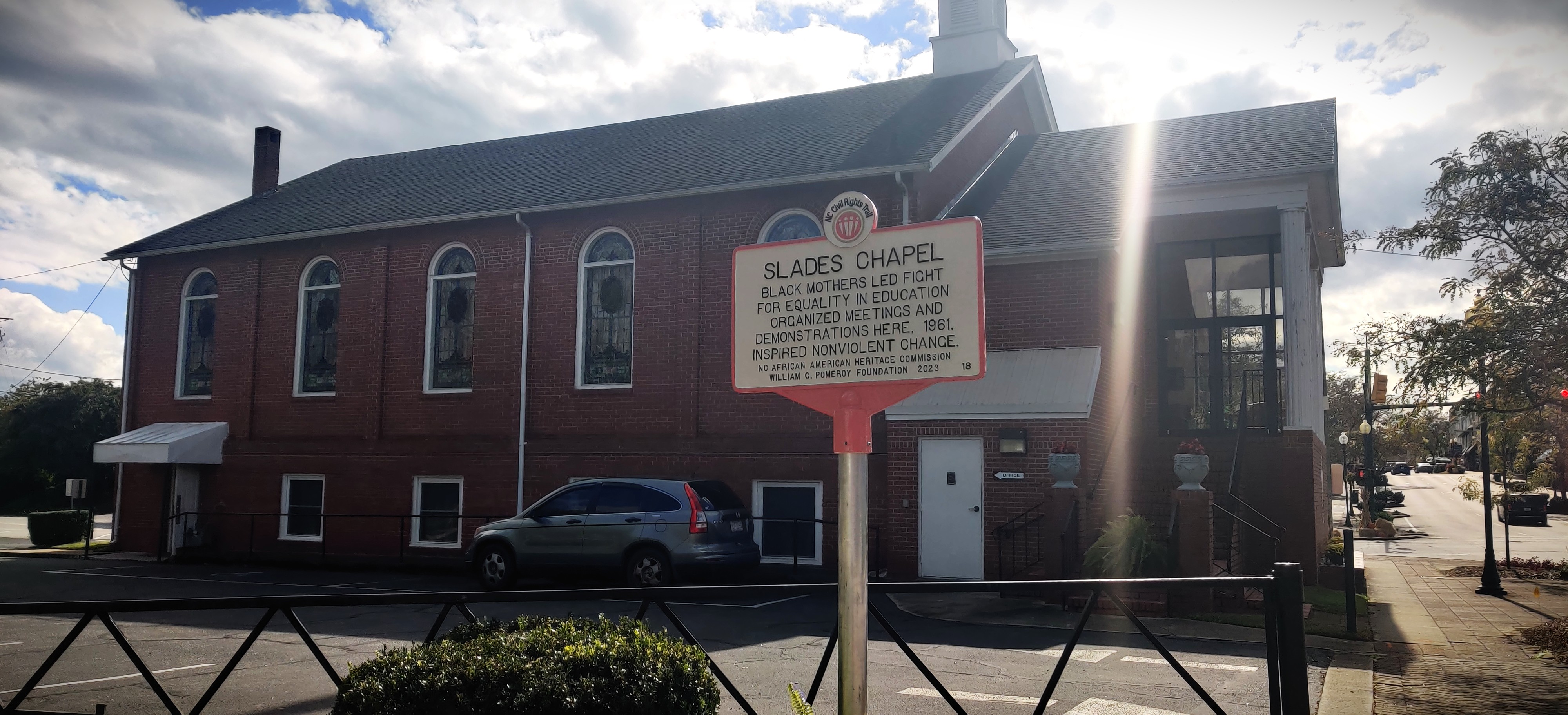 Photograph of historical marker honoring Seven Mothers. Marker has a white background with black lettering and red outline on the edges. Marker reads, 'Slades Chapel. Black mothers led fight for equality in education. Organized meetings and demonstrations here. 1961. Inspired nonviolent change. N.C. African American Heritage Commission. William C. Pomeroy Foundation 2023. Slades Chapel AME Zion Church is visible in the background.